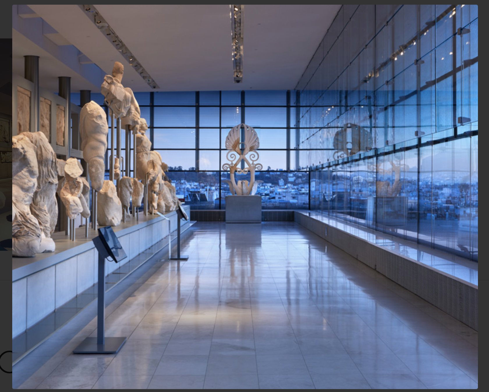 A photo of the national archaeological museum. It is taken early in the evening therefore a blue light from the sky comes from the large windows. On the left there is a display of marble statues